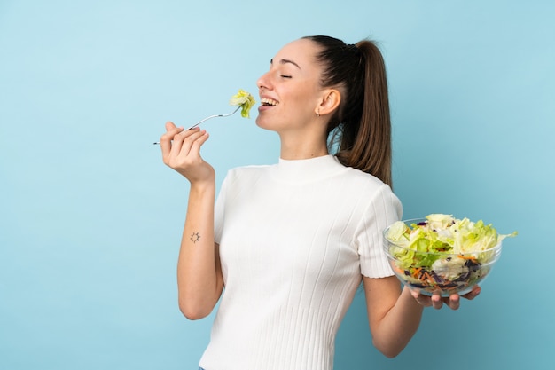 Junge brünette Frau, die einen Salat über isolierter blauer Wand hält