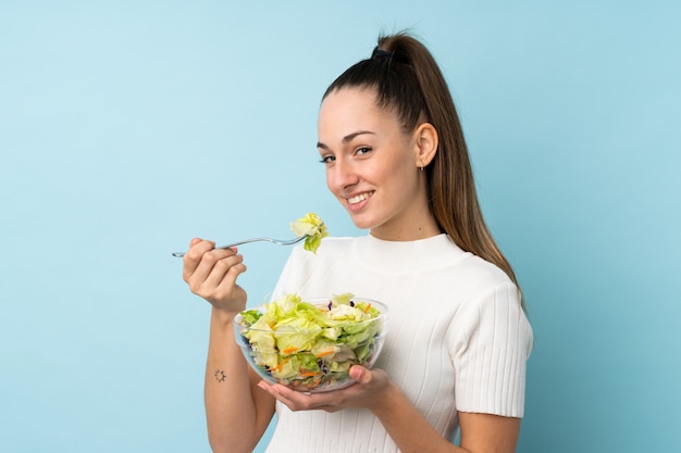 Junge brünette Frau, die einen Salat über isolierter blauer Wand hält