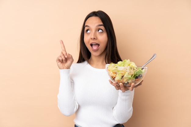 Junge brünette Frau, die einen Salat über isolierter Wand hält, die beabsichtigt, die Lösung zu realisieren, während ein Finger anhebt