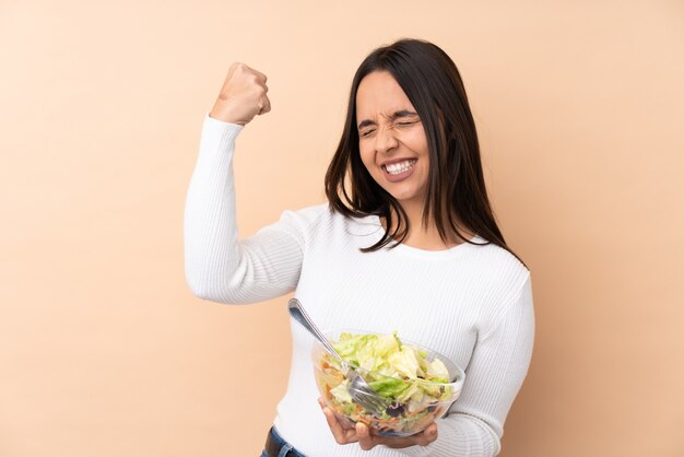 Junge brünette Frau, die einen Salat über isolierter Wand hält, die einen Sieg feiert