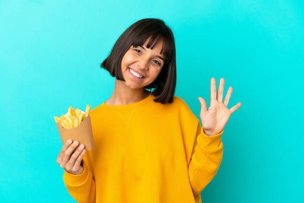 Junge brünette Frau, die gebratene Chips über isoliertem blauem Hintergrund hält und fünf mit den Fingern zählt