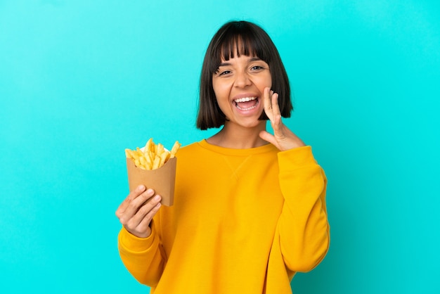 Junge brünette Frau, die gebratene Chips über isoliertem blauem Hintergrund hält und mit weit geöffnetem Mund schreit