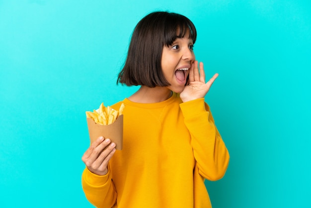 Junge brünette Frau, die gebratene Chips über isoliertem blauem Hintergrund hält und mit weit geöffnetem Mund zur Seite schreit