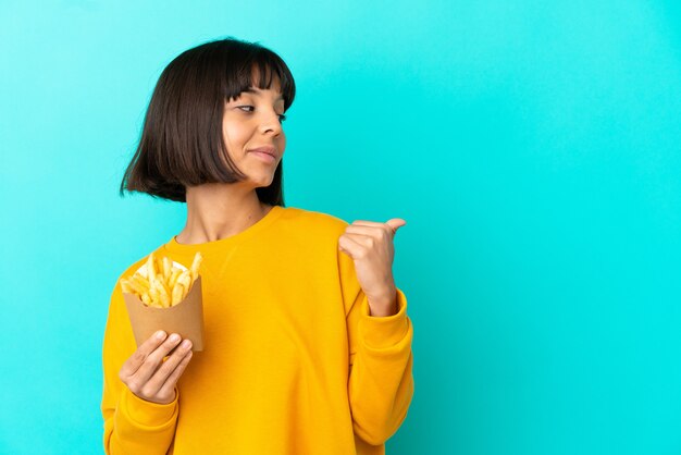 Junge brünette Frau, die gebratene Chips über isolierter blauer Wand hält und auf die Seite zeigt, um ein Produkt zu präsentieren