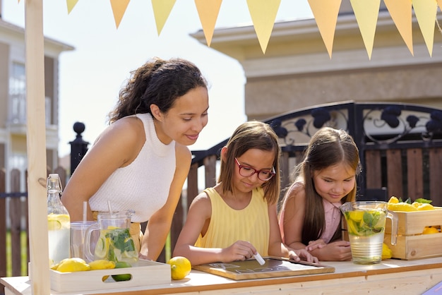 Junge brünette Frau, die ihre Tochter betrachtet, die auf Kündigung auf kleinem Brett mit Stück Kreide beim Verkauf von Limonade am sonnigen Tag zeigt