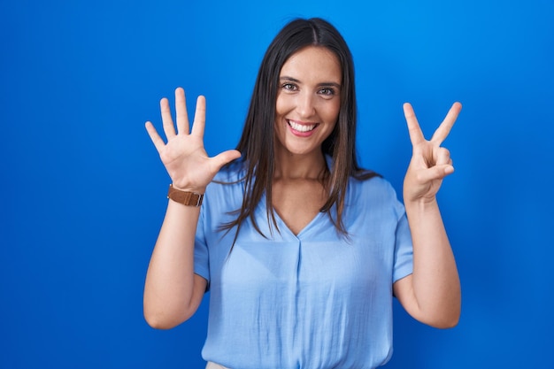 Junge brünette Frau, die vor blauem Hintergrund steht und mit den Fingern Nummer sieben nach oben zeigt, während sie selbstbewusst und glücklich lächelt.