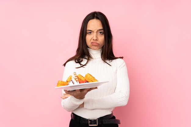 Junge brünette Frau, die Waffeln über isolierte rosa Wand hält, die sich verärgert fühlt