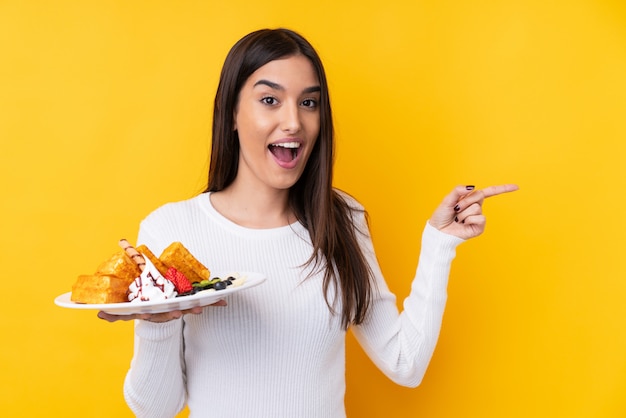 Junge brünette Frau, die Waffeln über isolierte Wand hält überrascht und Finger zur Seite zeigt