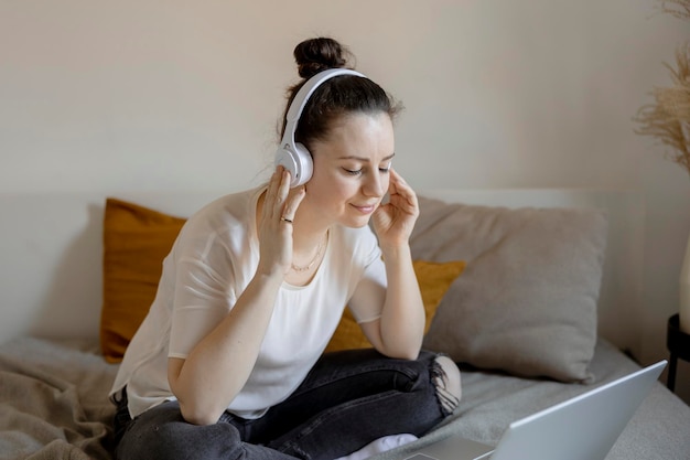 Junge brünette Frau, die zu Hause mit Freizeitkleidung auf dem Bett sitzt und Musik oder Hörbuch hört Entspannendes Ausruhen Zeit mit sich selbst psychische Gesundheit Mädchen träumt