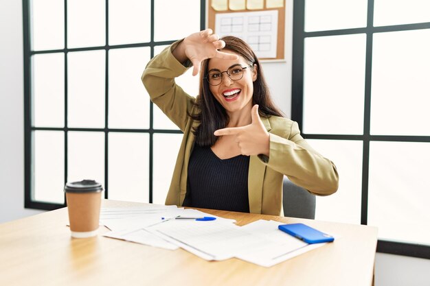 Foto junge brünette frau im business-stil sitzt am schreibtisch im büro und lächelt und macht rahmen mit händen und fingern mit glücklichem gesicht, kreativität und fotografie-konzept