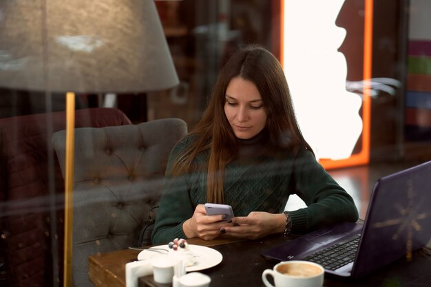 Junge brünette Frau im Café mit Laptop kommuniziert per Smartphone