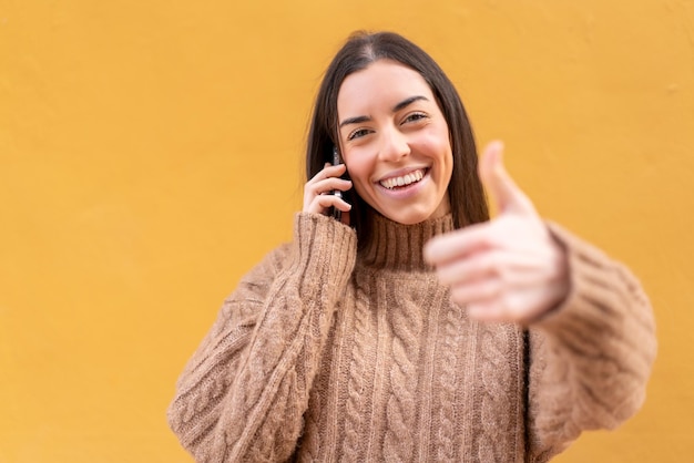 Junge brünette Frau im Freien, die ein Gespräch mit dem Handy führt, während sie Daumen hoch macht