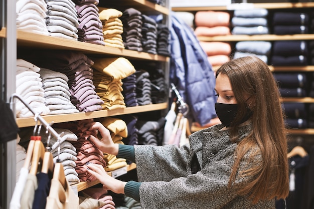 Junge brünette Frau in der medizinischen Maske wählt Kleidung im Laden und macht Einkäufe