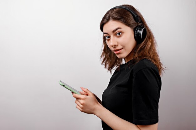 Junge brünette Frau in einem schwarzen T-Shirt auf Weiß hört Musik mit einem Telefon isoliert