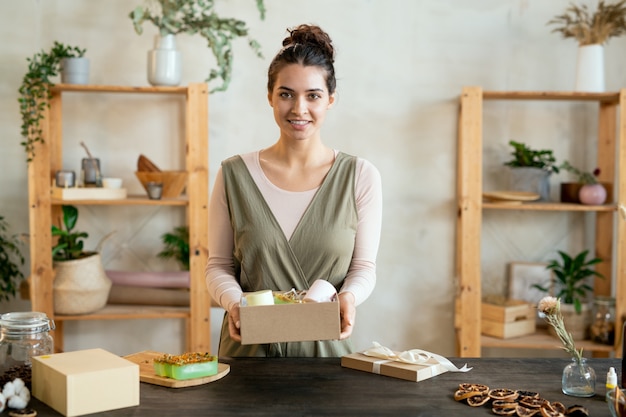 Junge brünette Frau in intelligenter Freizeitkleidung, die eine offene Geschenkbox aus Karton mit handgemachtem Geschenk hält, während sie vor der Kamera steht