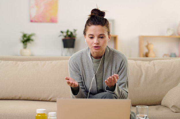 Junge brünette Frau in Kopfhörern, die dem Online-Arzt ihr Problem erklärt, während sie auf der Couch vor dem Laptop in der häuslichen Umgebung sitzt