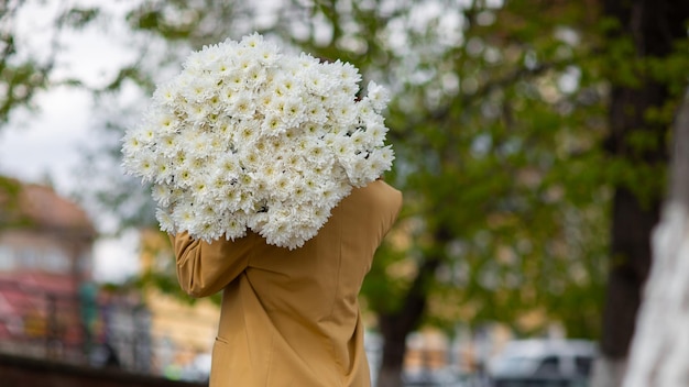 Junge brünette Frau in modernem beigefarbenem Outfit mit einem Strauß Chrysantemblumen, die in der sonnigen europäischen Stadtstraße spazieren gehen Rückansicht Ästhetik