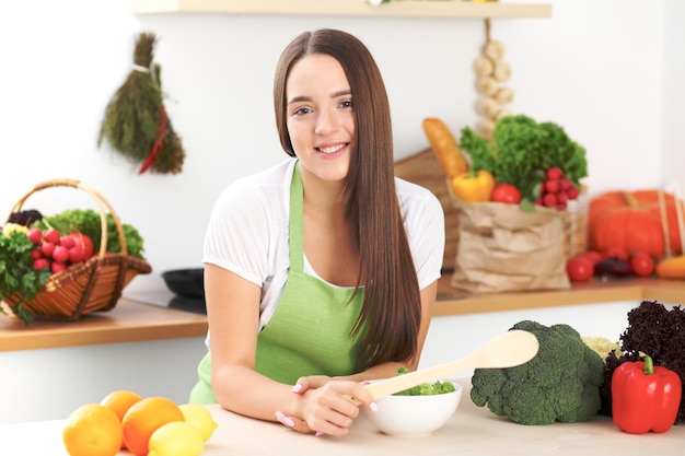 Junge brünette Frau kocht oder isst frischen Salat in der Küche Hausfrau mit Holzlöffel in der rechten Hand Lebensmittel- und Gesundheitskonzept