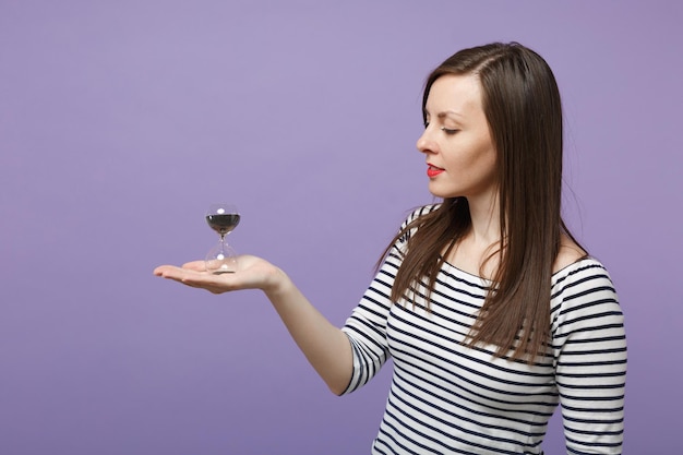 Junge brünette Frau Mädchen in lässig gestreifter Kleidung posiert isoliert auf violettem Hintergrund Studioportrait. Menschen aufrichtige Emotionen Lifestyle-Konzept. Mock up Copy Space Halten Sie Sanduhr schwarzen Sand