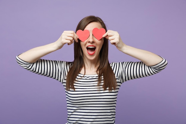 Junge brünette Frau Mädchen in lässig gestreifter Kleidung posiert isoliert auf violettem Hintergrund Studioportrait. Menschen aufrichtige Emotionen Lifestyle-Konzept Mock up Copy Space Halten Sie zwei kleine rote Herzen