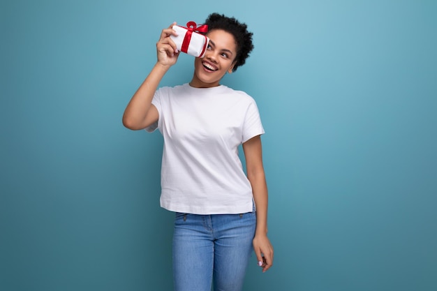 Junge brünette Frau mit Afro-Haaren hält ein Geschenk mit einer Schleife auf blauem Hintergrund mit Kopie