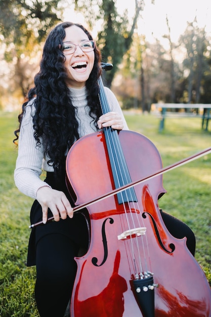 Junge brünette Frau mit Brille, die bei Sonnenuntergang im Park Cello spielt, auf grünem Gras. Vertikal