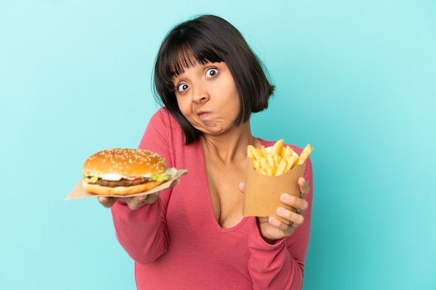 Junge brünette Frau mit Burger und gebratenen Pommes über isoliertem blauem Hintergrund