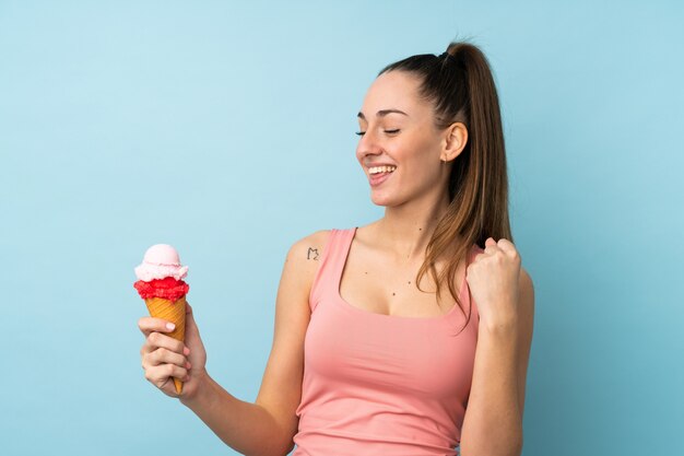 Junge brünette Frau mit einem Kornetteis über isolierter blauer Wand, die einen Sieg feiert