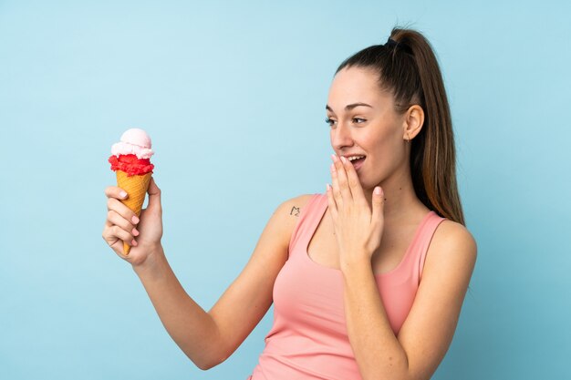 Junge brünette Frau mit einem Kornetteis über isolierter blauer Wand mit Überraschung und schockiertem Gesichtsausdruck