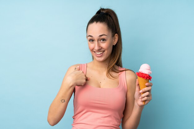 Junge brünette Frau mit einem Kornetteis über isolierter blauer Wand mit überraschendem Gesichtsausdruck