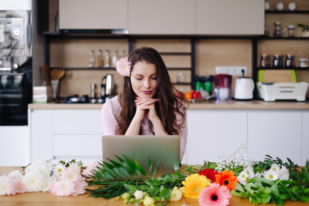 Junge brünette Frau mit langen gewellten Haaren, die am Laptop mit Blumen auf dem Schreibtisch mit Küche im Hintergrund arbeitet Talentierte Floristin entwickelt Online-Verkäufe und bereitet sich auf den Workshop vor