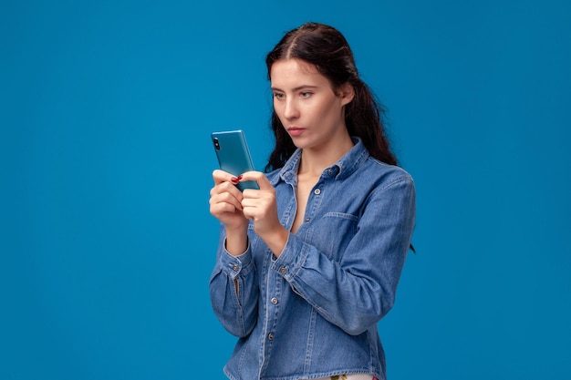 Junge brünette Frau posiert mit einem Smartphone auf blauem Hintergrund.