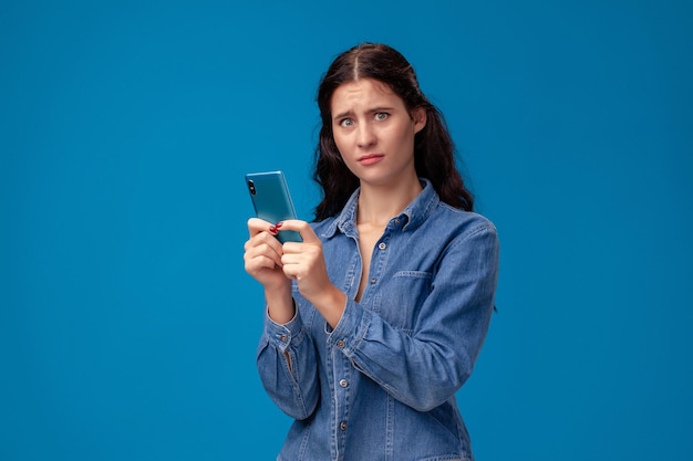 Junge brünette Frau posiert mit einem Smartphone auf blauem Hintergrund.