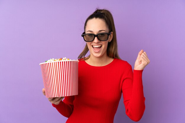 Junge Brünette Frau über isolierte lila Wand mit 3D-Brille und hält einen großen Eimer Popcorn