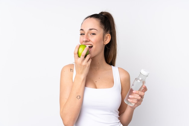 Junge brünette Frau über isolierte weiße Wand mit einer Flasche Wasser und Essen eines Apfels