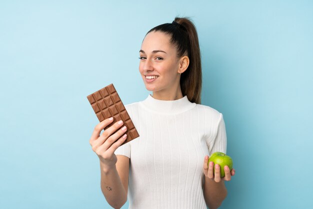 Junge brünette Frau über isolierter blauer Wand, die eine Schokoladentafel in einer Hand und einen Apfel in der anderen nimmt