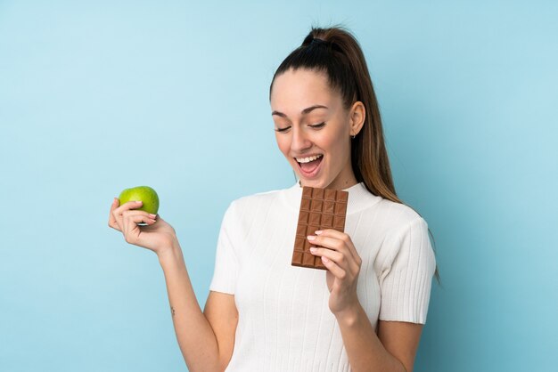 Junge brünette Frau über isolierter blauer Wand, die eine Schokoladentafel in einer Hand und einen Apfel in der anderen nimmt