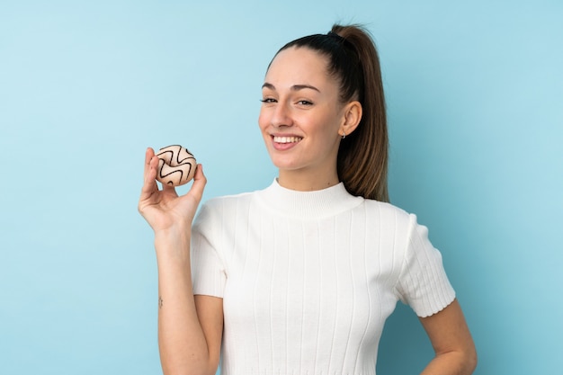 Junge brünette Frau über isolierter blauer Wand, die einen Donut und glücklich hält