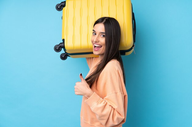 Junge brünette Frau über isolierter blauer Wand im Urlaub mit Reisekoffer und mit Daumen nach oben