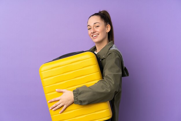 Junge brünette Frau über isolierter lila Wand im Urlaub mit Reisekoffer