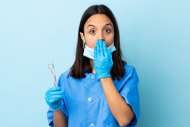 Junge Brünette gemischte Rasse Zahnarzt Frau, die Werkzeuge über Wand bedeckt Mund mit Hand hält