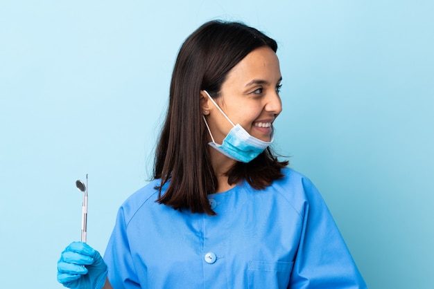 Junge Brünette gemischte Rasse Zahnarztfrau, die Werkzeuge über lokalisiertem Hintergrund hält, der zur Seite schaut und lächelt