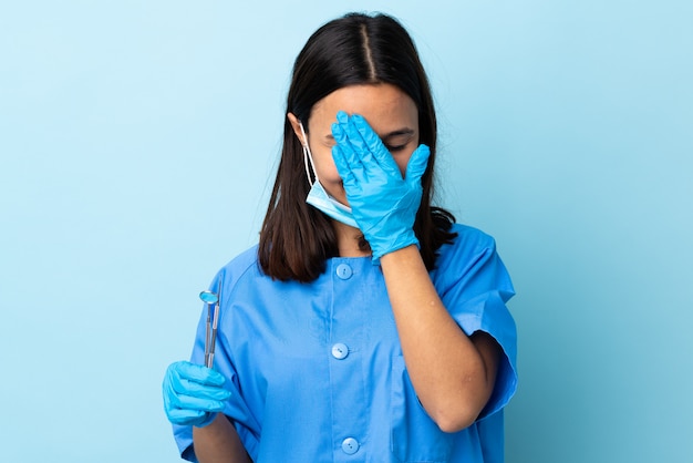 Junge brünette gemischte zahnärztin, die werkzeuge über wand hält, die augen durch hände bedeckt