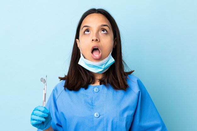 Junge brünette gemischte Zahnarztfrau, die Werkzeuge über isolierter Wand hält, die oben und mit überraschtem Ausdruck schaut