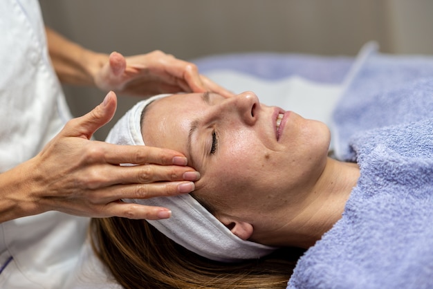 Junge Brünette genießt die reinigende Gesichtsmassage mit geschlossenen Augen auf einem Tisch liegend.