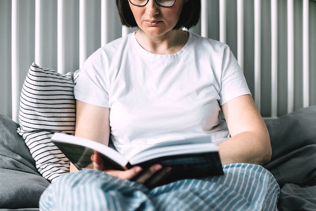 Junge Brünette im weißen Hemd liest Buch auf dem Bett zu Hause. Konzept des gemütlichen, komfortablen Zuhauses. Lifestyle, authentischer Moment