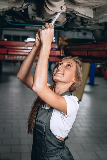 Junge Brünette in schwarzer Arbeitskleidung und weißem T-Shirt schraubt die Mutter mit einem Schraubenschlüssel unter dem Auto ab und lächelt. Mechanikerin arbeitet unter der Unterseite eines Autos