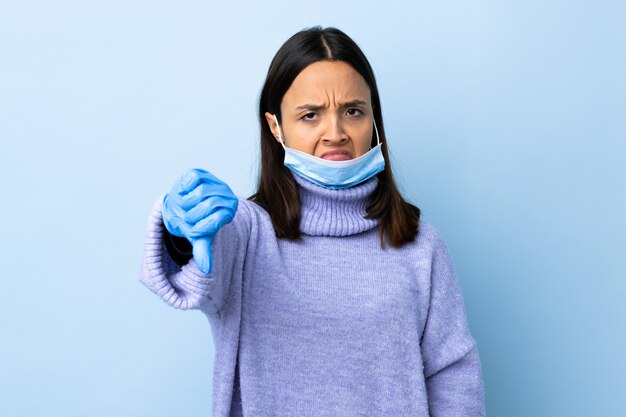 Junge brünette Mischlingsfrau, die vor dem Coronavirus mit einer Maske und Handschuhen über isolierter blauer Wand schützt, die Daumen unten mit negativem Ausdruck zeigt