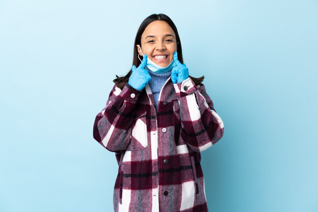 Junge brünette Mischlingsfrau, die vor dem Coronavirus mit einer Maske und Handschuhen über isolierter blauer Wand schützt, die mit einem glücklichen und angenehmen Ausdruck lächelt