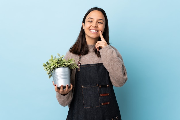 Junge brünette Mischrassenfrau, die eine Pflanze über isoliertem blauem Hintergrund hält, der mit einem glücklichen und angenehmen Ausdruck lächelt